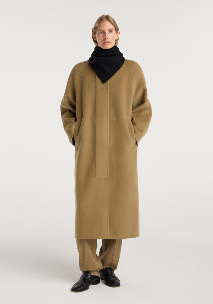 Person wearing a long camel coat and black shoes, styled with a Small knitted triangle scarf | black wrapped around the neck, against a plain light background.
