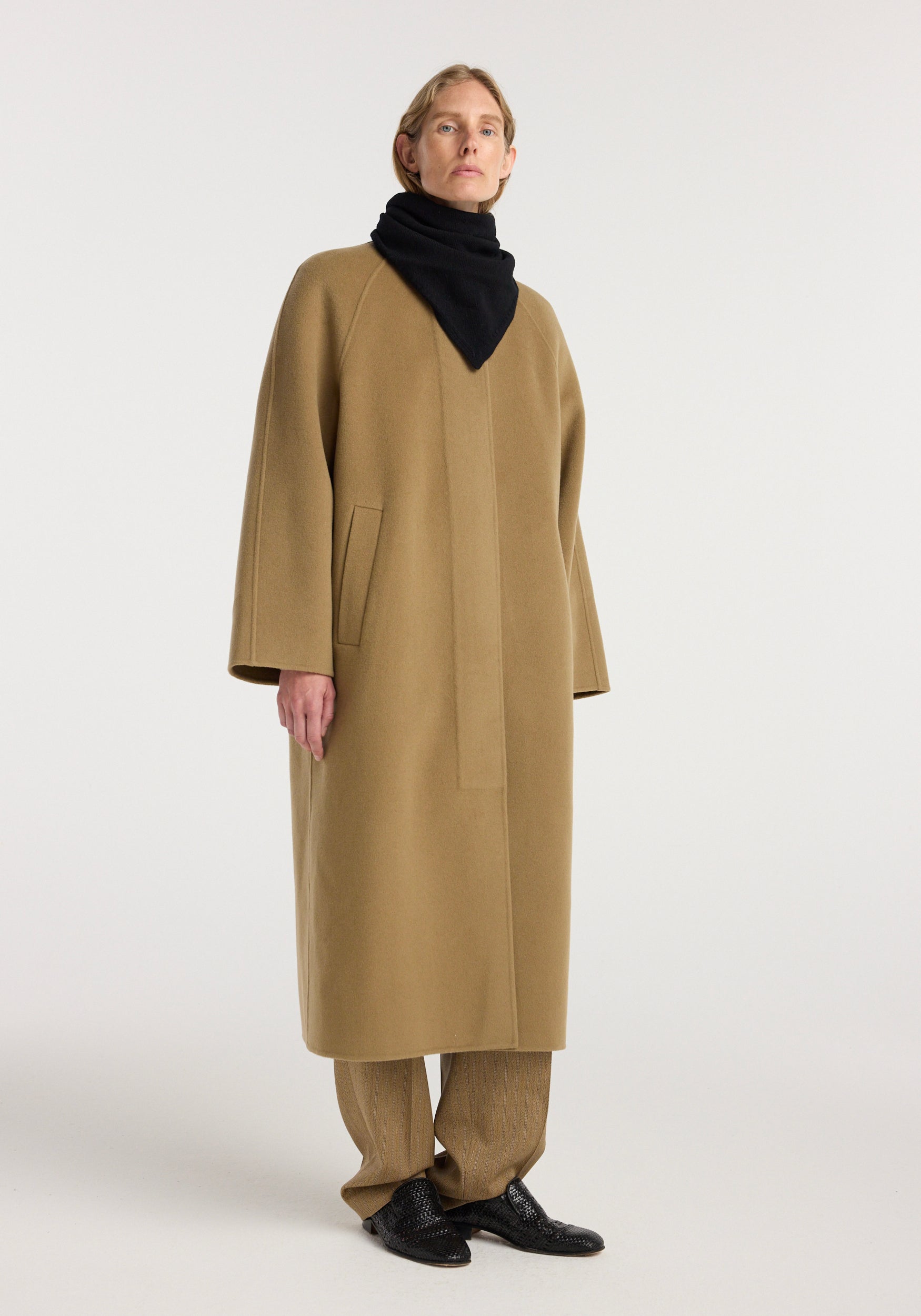 A person wearing a long camel-colored coat and brown pants, styled with a Small knitted triangle scarf | black wrapped around the neck, standing against a plain light background.