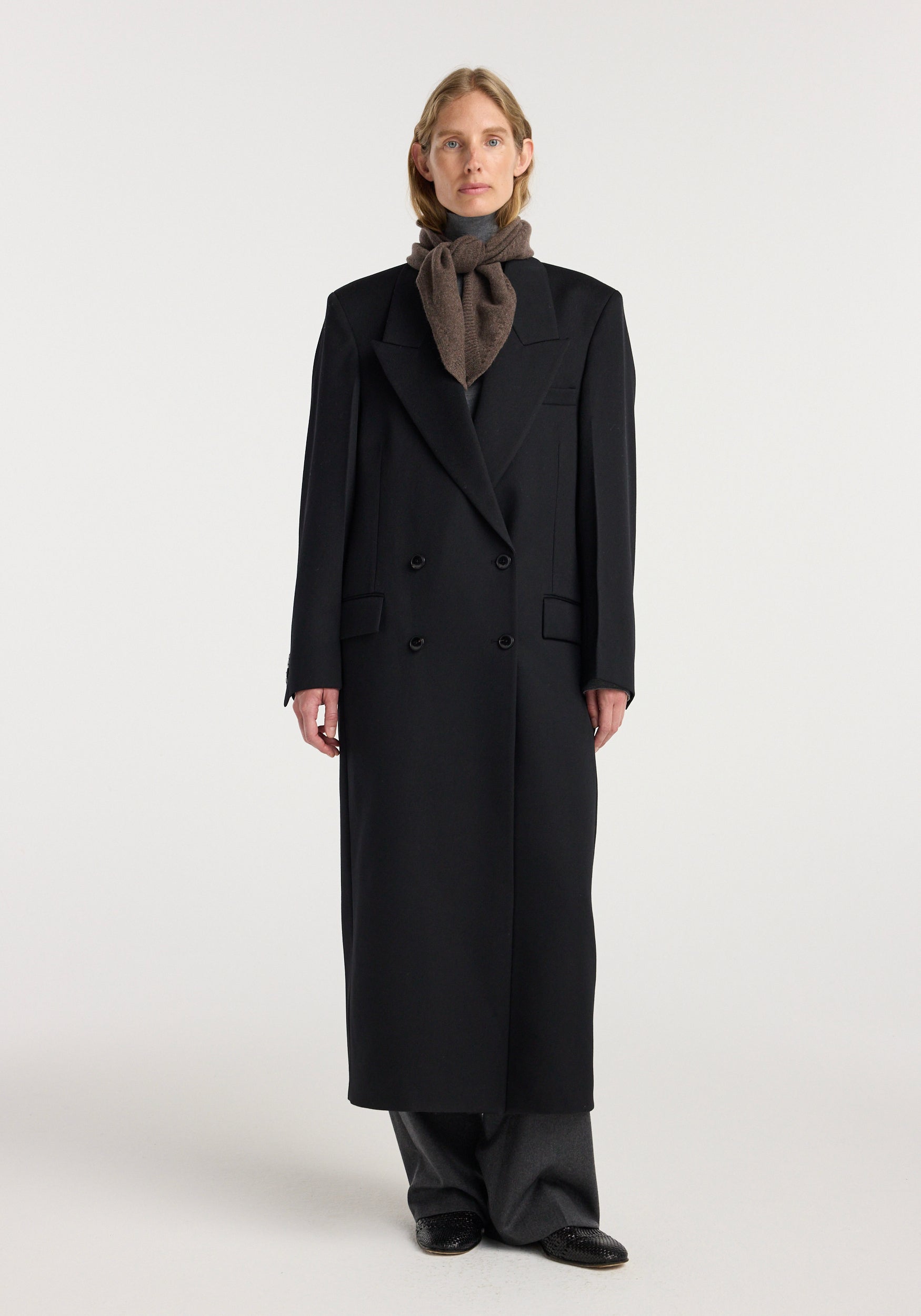 Model wearing a Small knitted triangle scarf | brown melange tied neatly around the neck paired with a long black coat and gray pants.