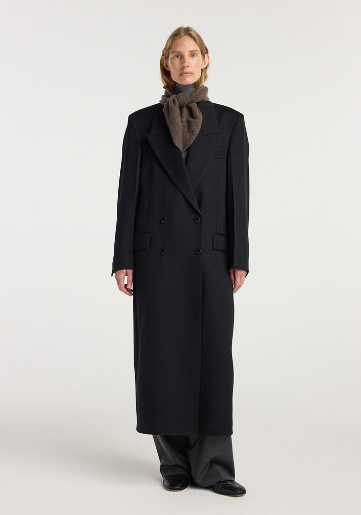 Model wearing a Small knitted triangle scarf | brown melange tied neatly around the neck paired with a long black coat and gray pants.