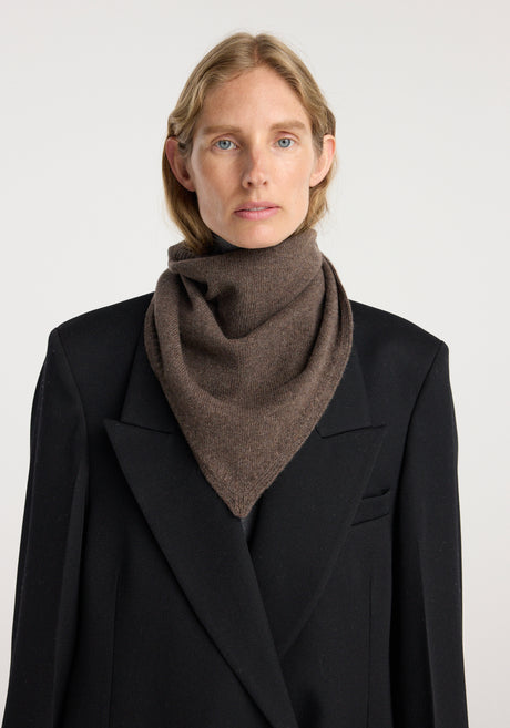 Model wearing a Small knitted triangle scarf | brown melange tied neatly around the neck paired with a long black coat and gray pants.