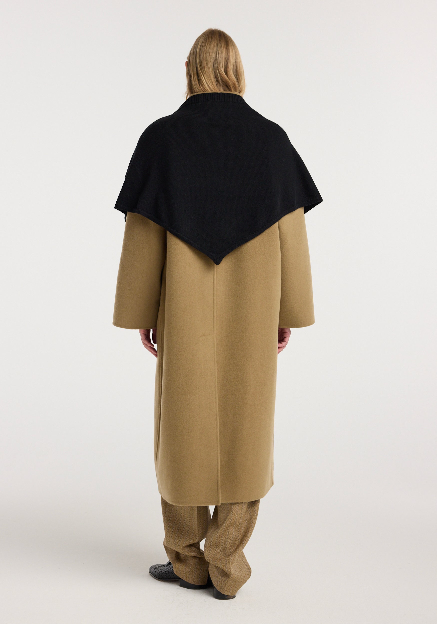 Woman seen from behind wearing a large knitted triangle scarf | black draped over a long camel coat and matching wide-leg pants against a plain background.