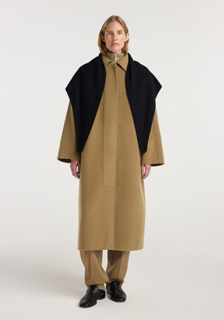 Model wearing a large knitted triangle scarf | black draped over shoulders, paired with a long camel coat, matching pants, and black shoes, standing against a plain light background.