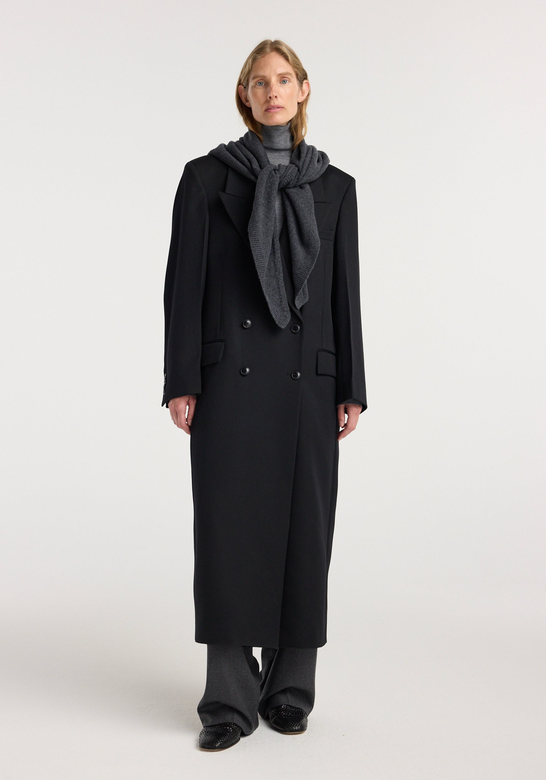 Model wearing a large knitted triangle scarf | anthracite melange, styled draped and knotted over a black long coat with dark gray trousers and black shoes against a plain background.