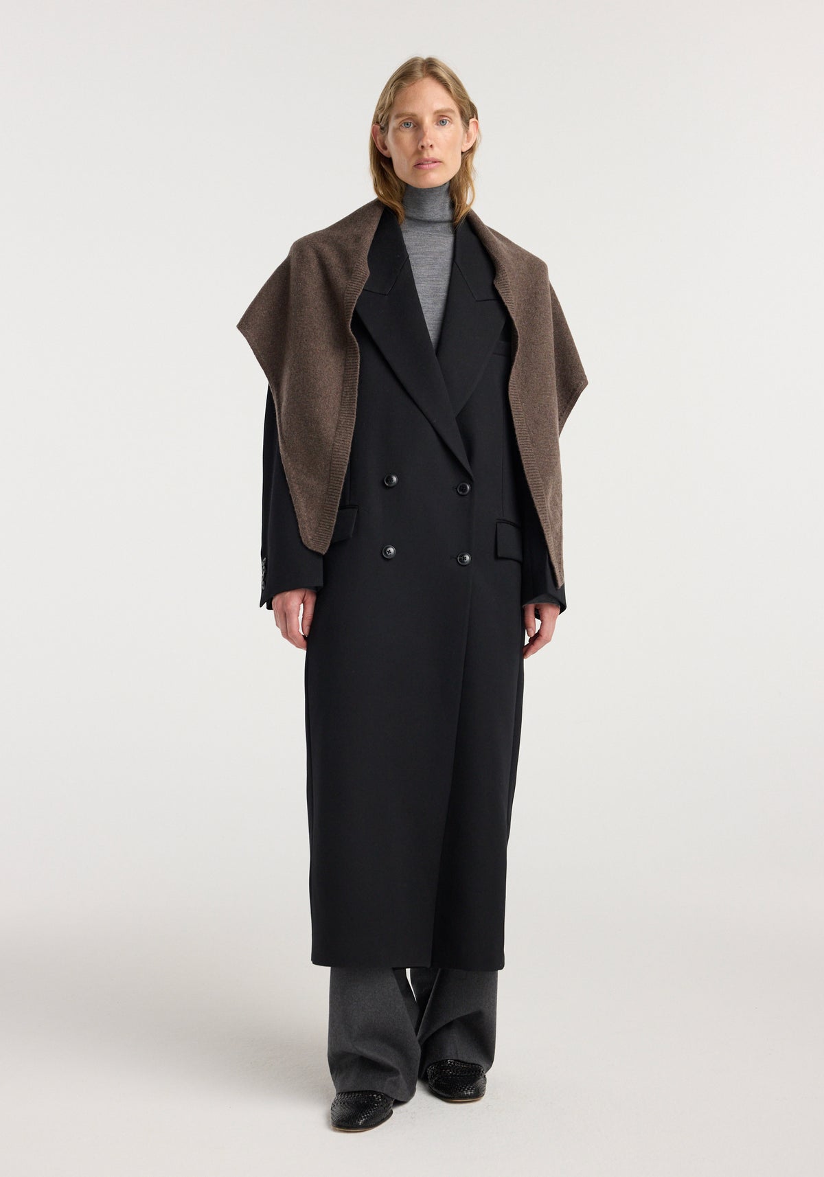 Woman wearing a black long coat over a gray turtleneck and trousers, with a Large knitted triangle scarf | brown melange draped over her shoulders.