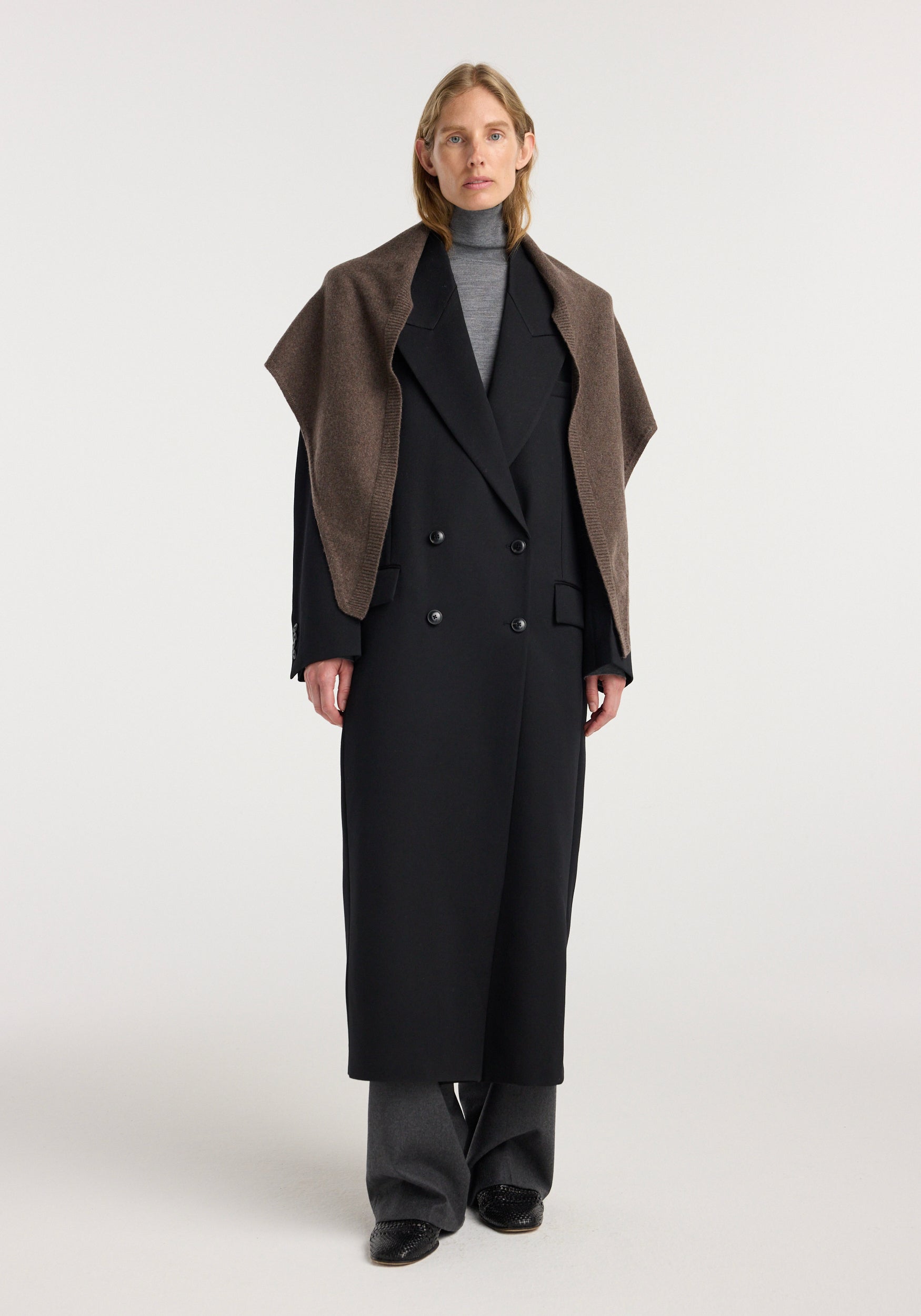 Woman wearing a black long coat over a gray turtleneck and trousers, with a Large knitted triangle scarf | brown melange draped over her shoulders.
