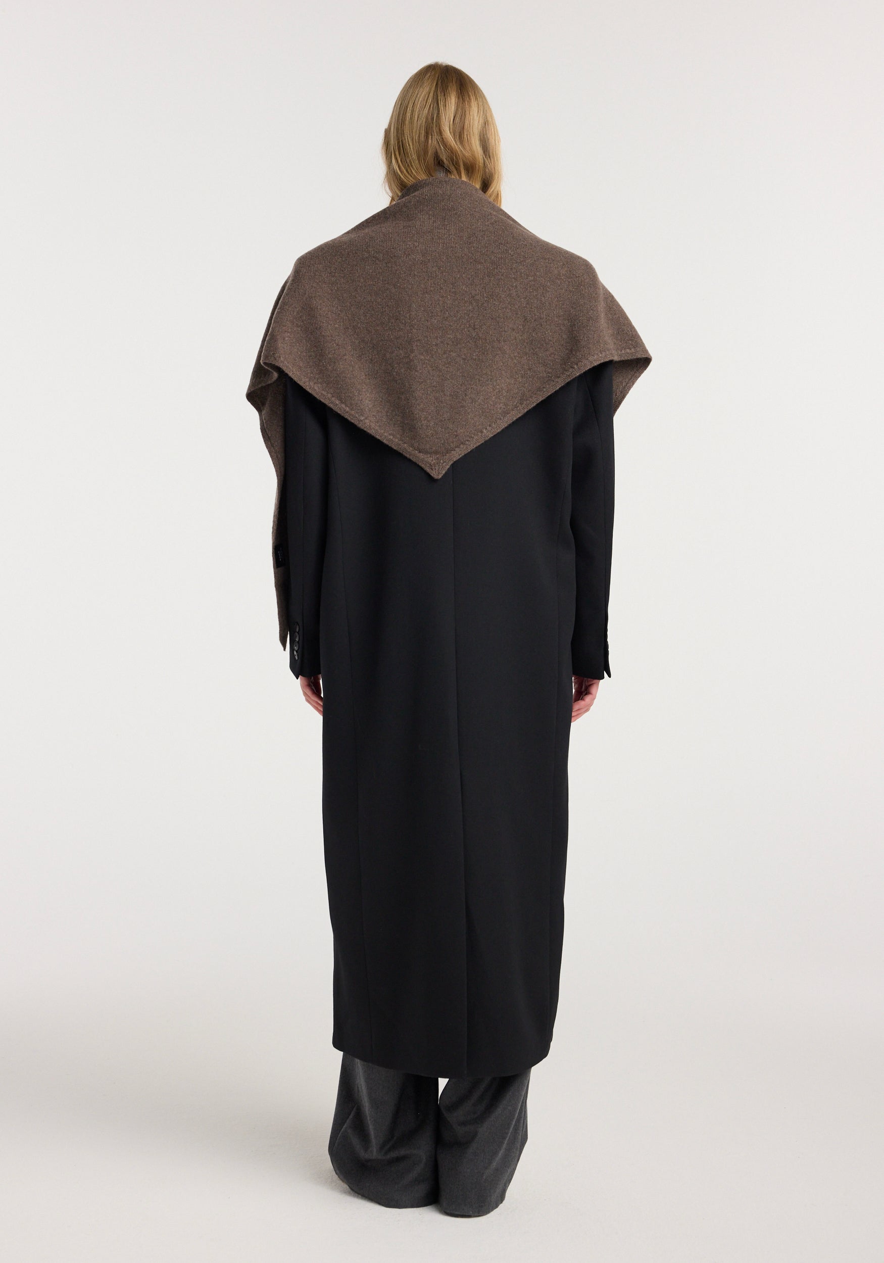 Back view of a person wearing a Large knitted triangle scarf | brown melange draped over a long black coat against a plain white background.