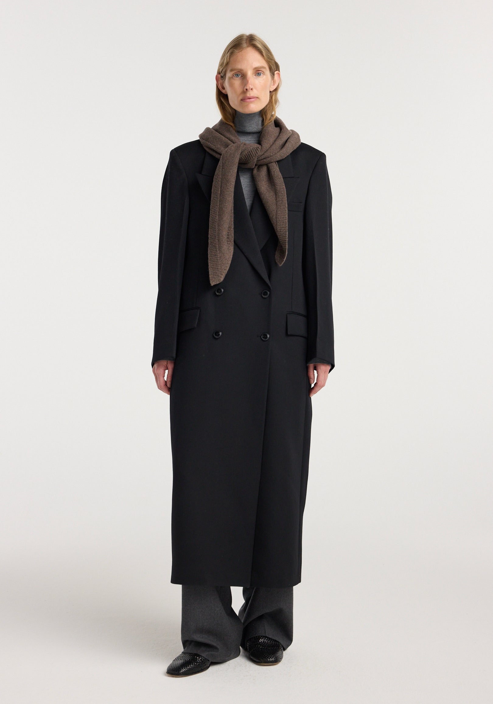 Woman wearing a large knitted triangle scarf | brown melange wrapped around her neck with a long black double-breasted coat, gray turtleneck, gray pants, and black shoes against a plain background.