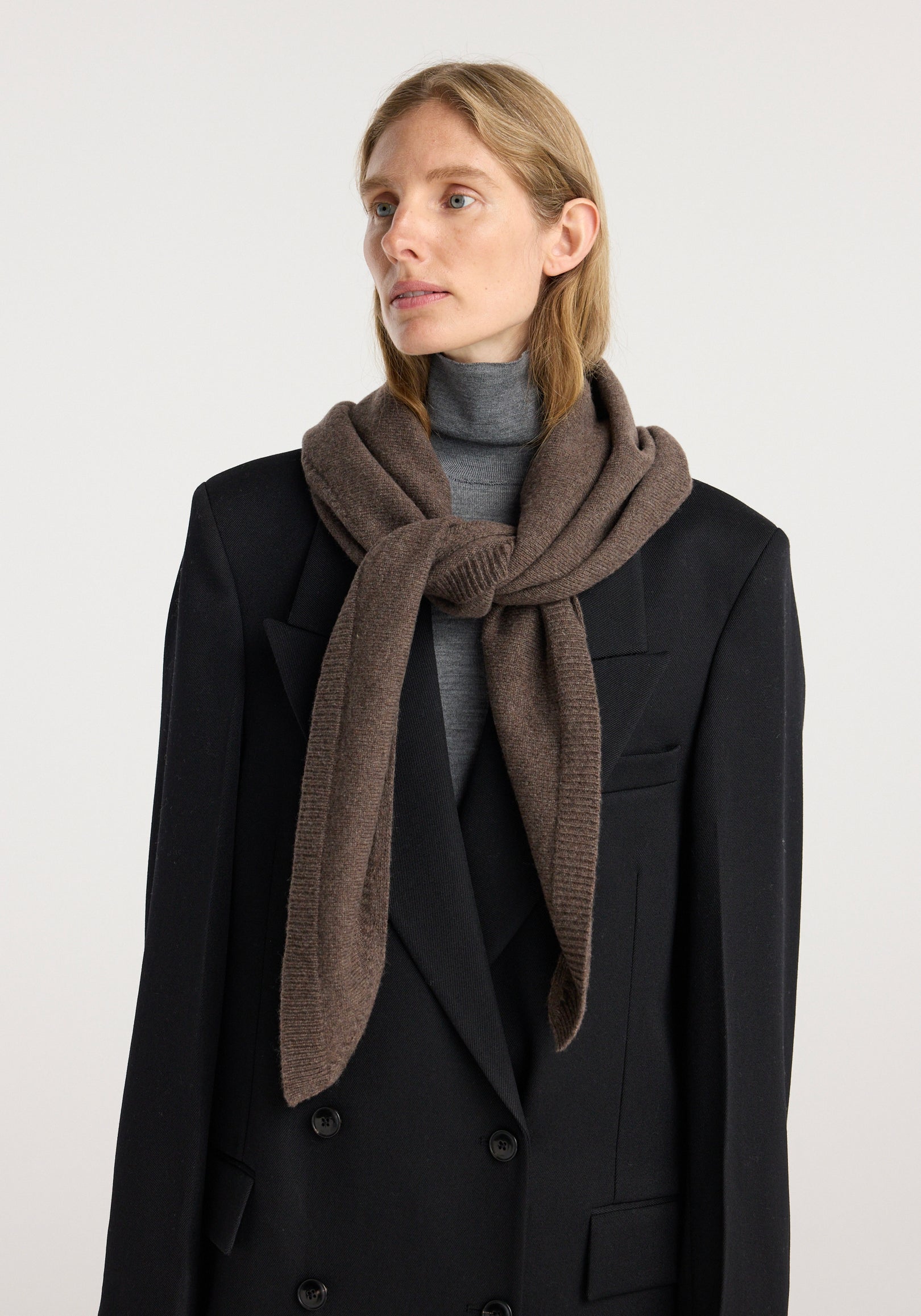 Woman wearing a large knitted triangle scarf | brown melange, styled over a black double-breasted coat and grey turtleneck sweater against a plain background.