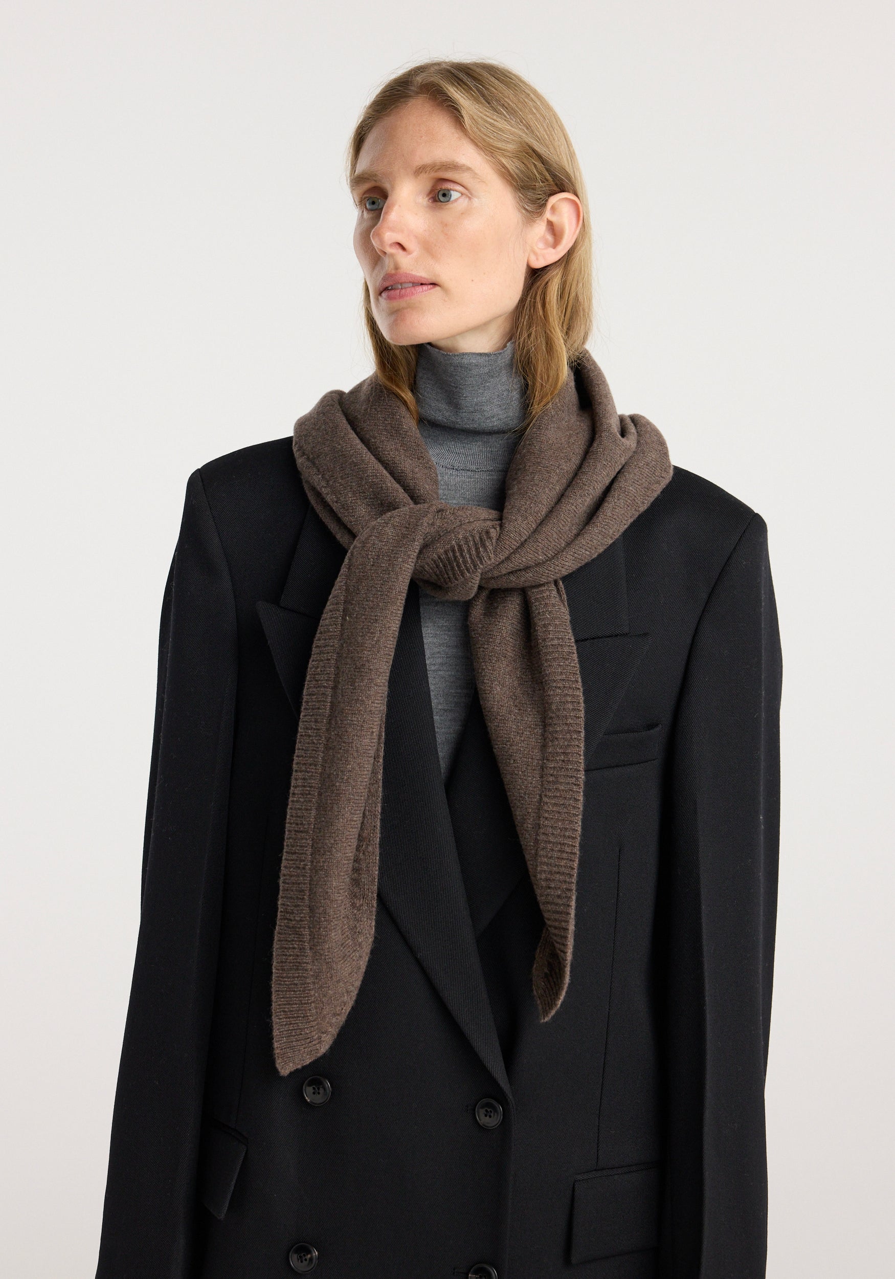 Woman wearing a large knitted triangle scarf | brown melange, styled over a black double-breasted coat and grey turtleneck sweater against a plain background.