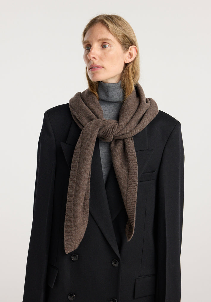 Woman wearing a large knitted triangle scarf | brown melange, styled over a black double-breasted coat and grey turtleneck sweater against a plain background.