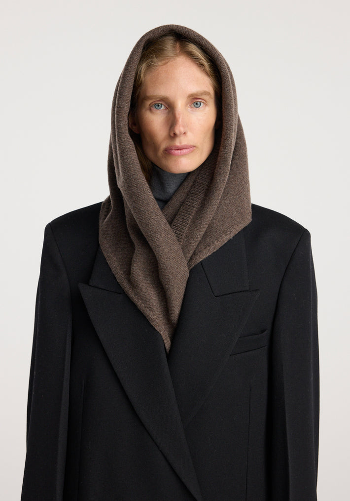 Woman wearing a large knitted triangle scarf | brown melange styled as a hood over her head, paired with a black tailored coat against a neutral background.