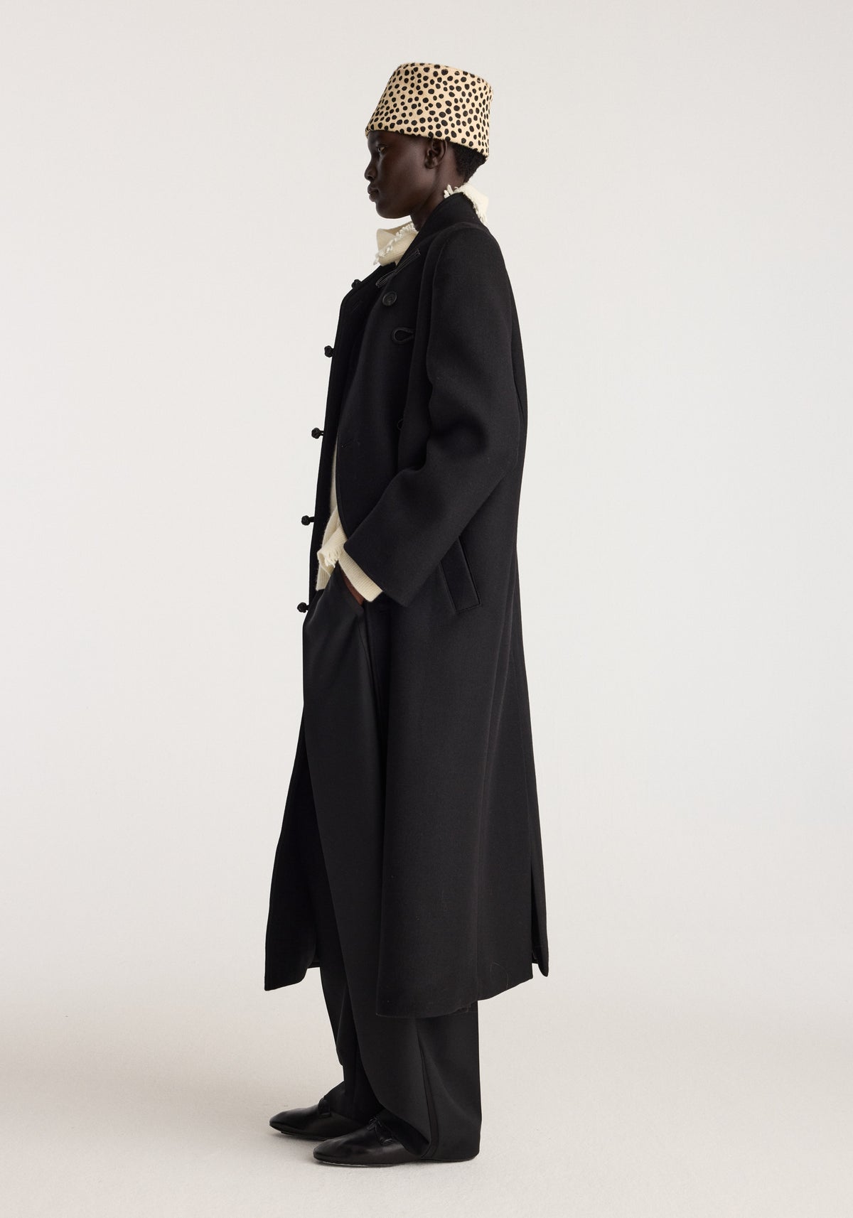 Cheetah leather hat | cheetah worn by a model in profile with a long black coat and trousers against a neutral studio background