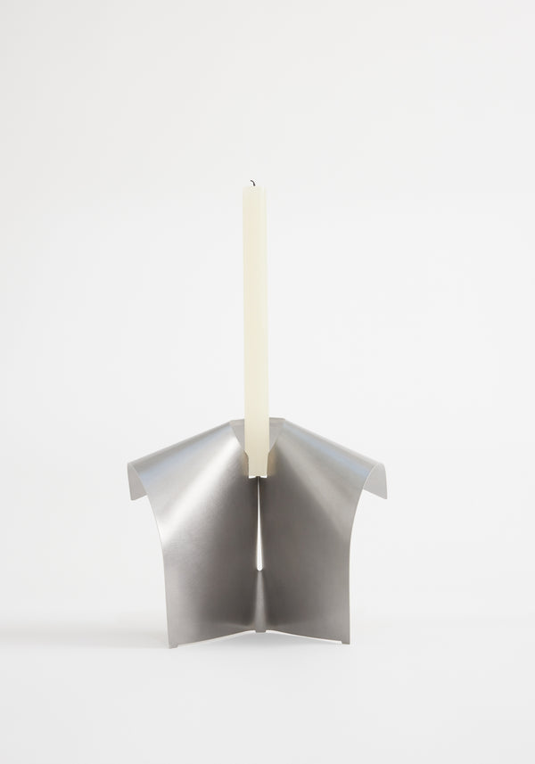 Stainless steel candle holder featuring a minimalist, tripod-like design from folded metal, securely holding an ivory taper candle. Designed by Paul Coenen, handcrafted in the Netherlands.
