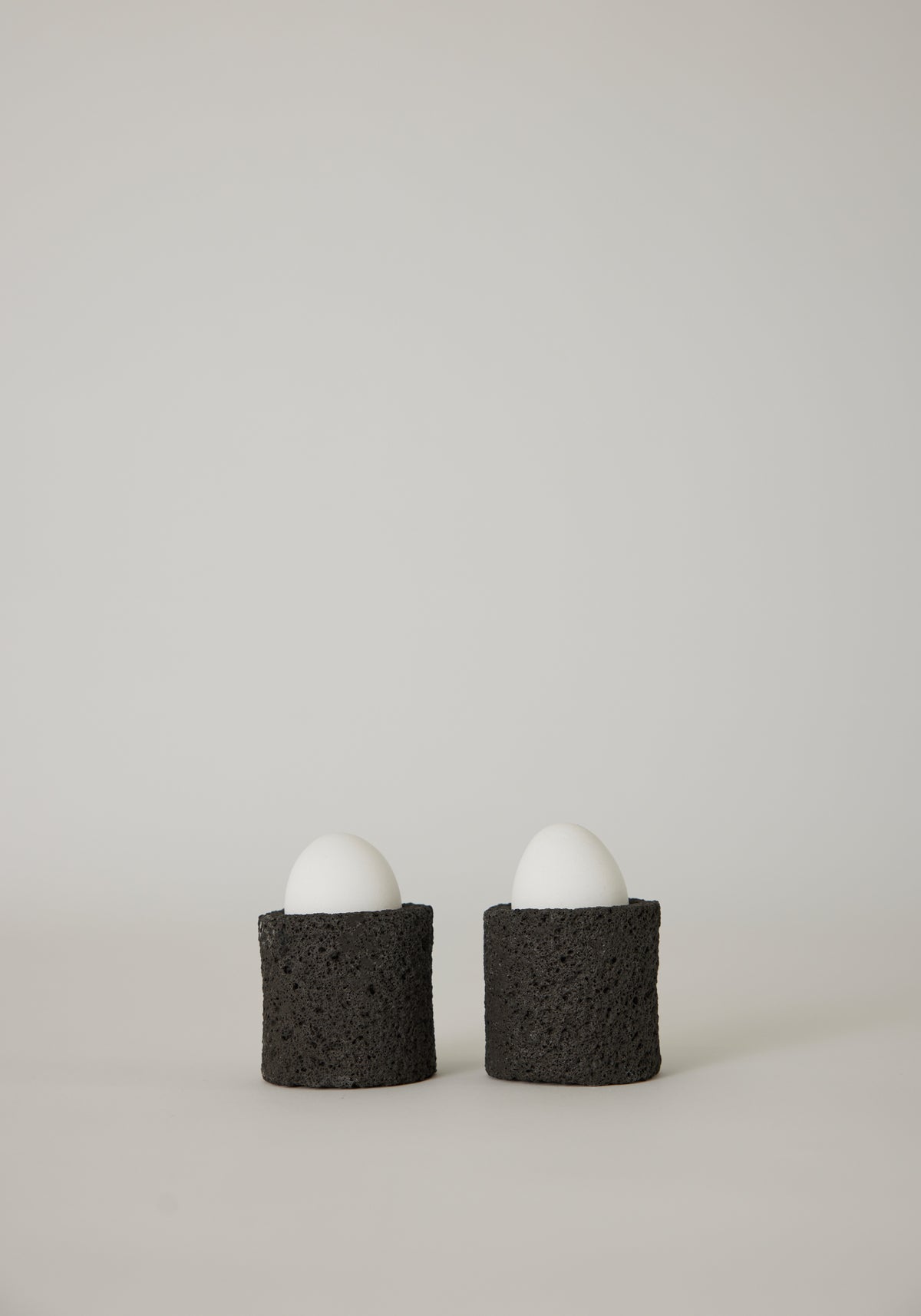Egg Cups: Two white eggs in handcrafted, dark-gray lava stone holders by Louis Crombag, showcasing a unique, textured design from the Netherlands.