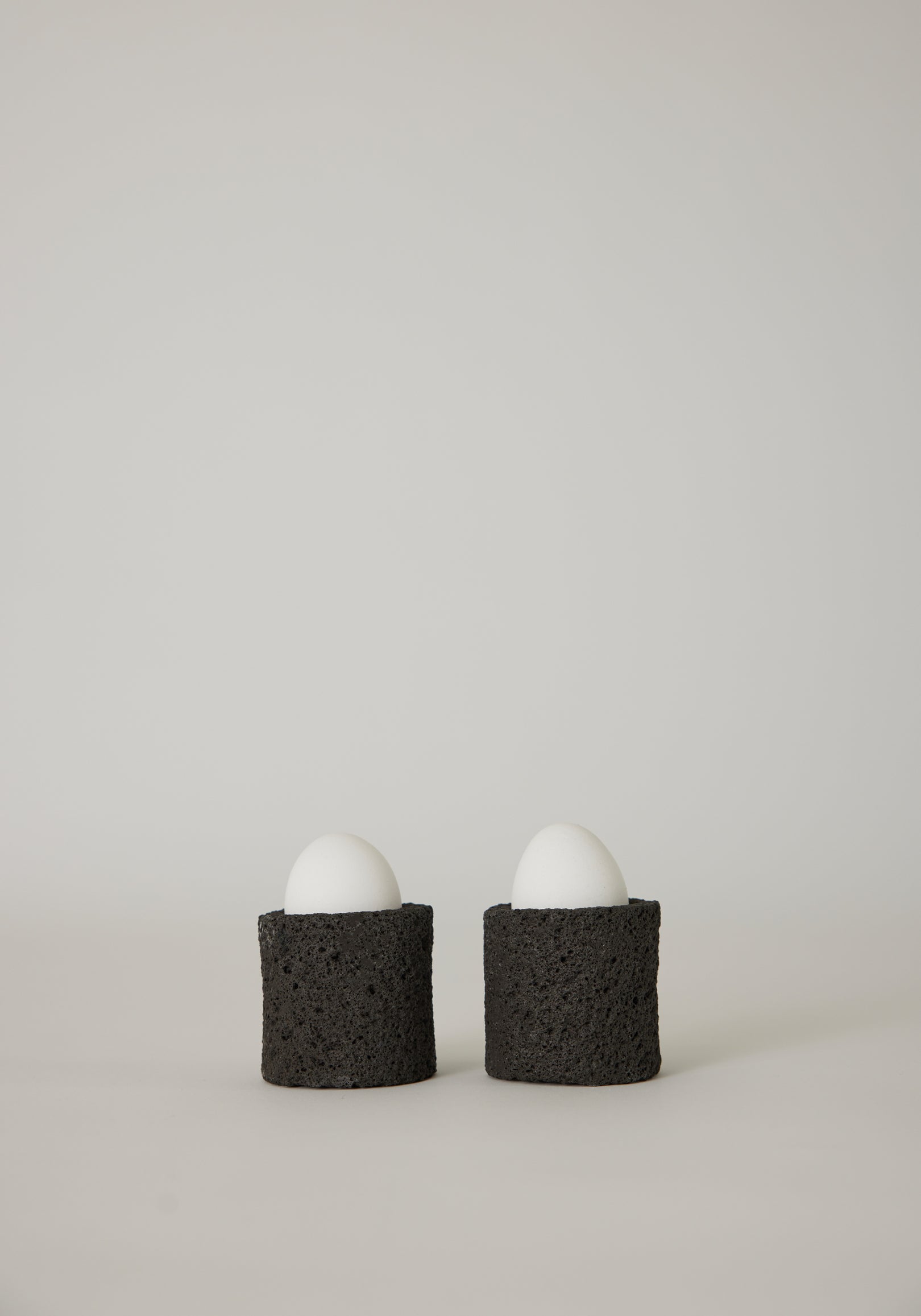 Egg Cups: Two white eggs in handcrafted, dark-gray lava stone holders by Louis Crombag, showcasing a unique, textured design from the Netherlands.