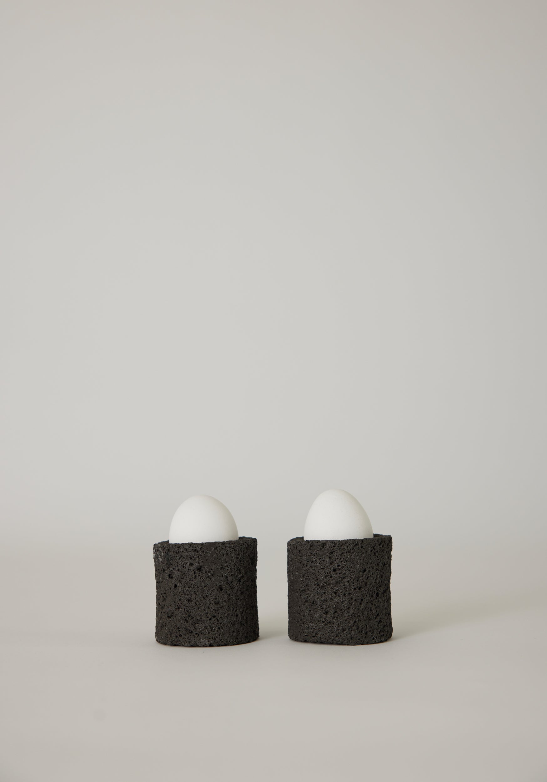 Egg Cups: Two white eggs in handcrafted, dark-gray lava stone holders by Louis Crombag, showcasing a unique, textured design from the Netherlands.