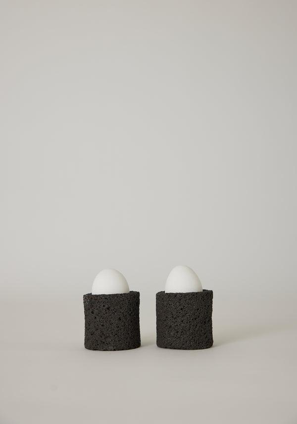 Egg Cups: Two white eggs in handcrafted, dark-gray lava stone holders by Louis Crombag, showcasing a unique, textured design from the Netherlands.