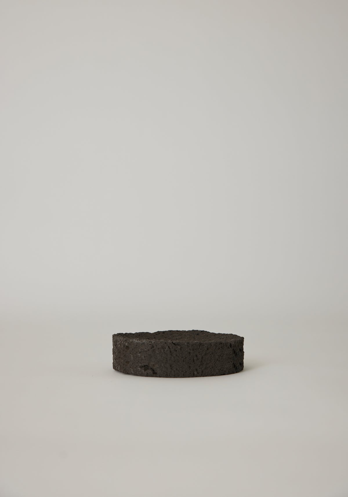 Round Tray made from black, porous lava stone, handcrafted in the Netherlands by Louis Crombag, featuring a flat top and carved indent, measuring 2 cm by 13.5 cm.