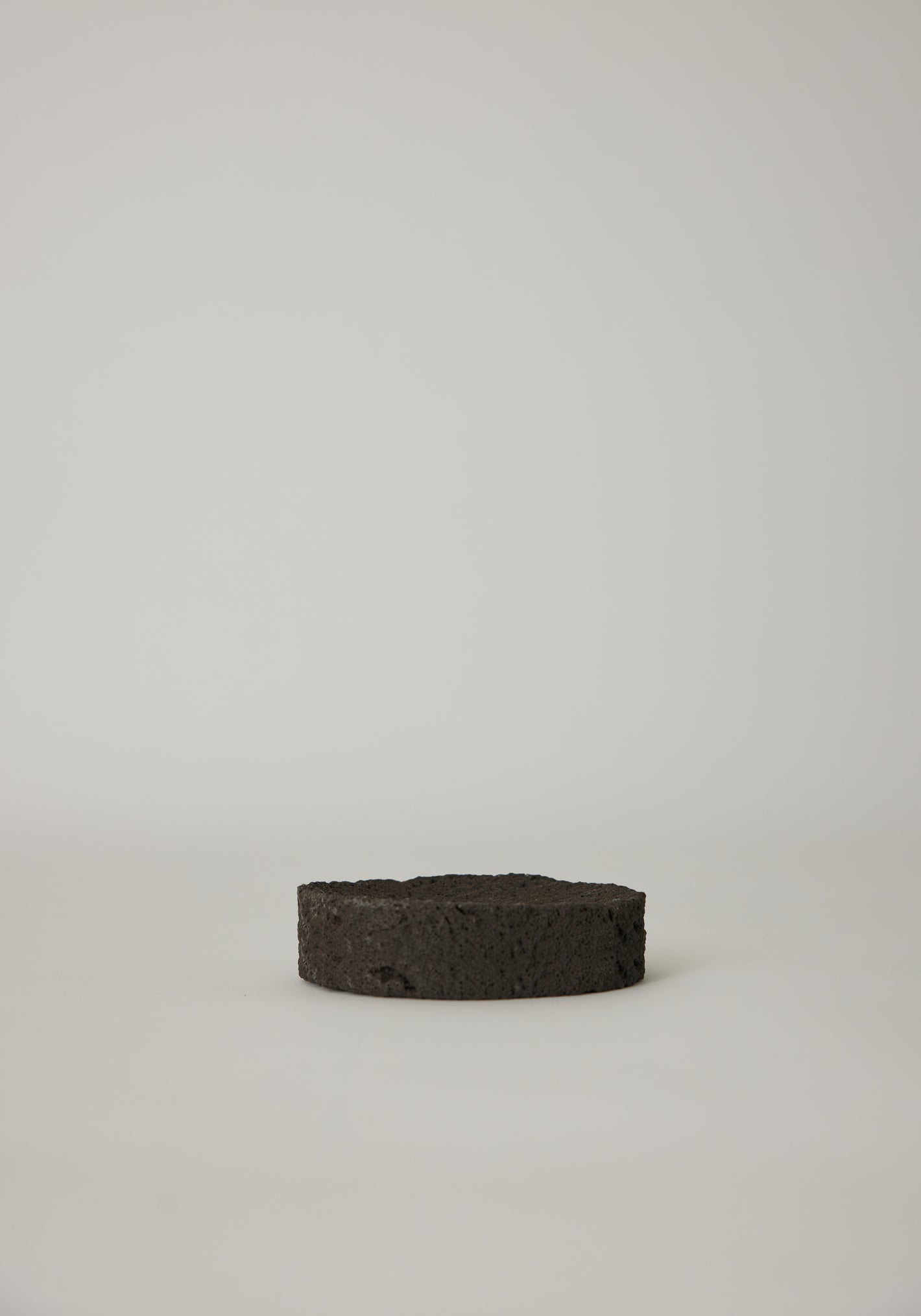 Round Tray made from black, porous lava stone, handcrafted in the Netherlands by Louis Crombag, featuring a flat top and carved indent, measuring 2 cm by 13.5 cm.