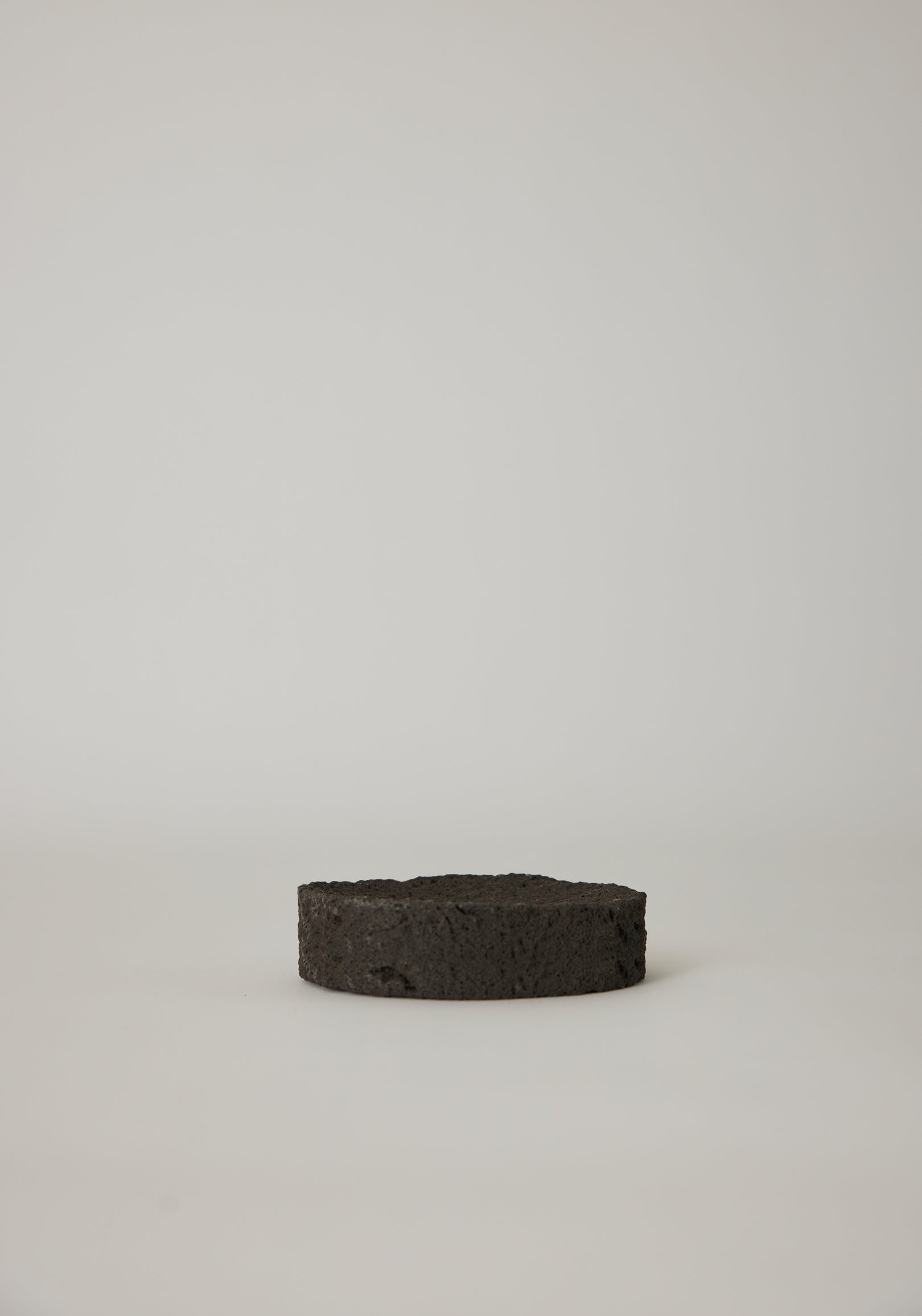 Round Tray made from black, porous lava stone, handcrafted in the Netherlands by Louis Crombag, featuring a flat top and carved indent, measuring 2 cm by 13.5 cm.