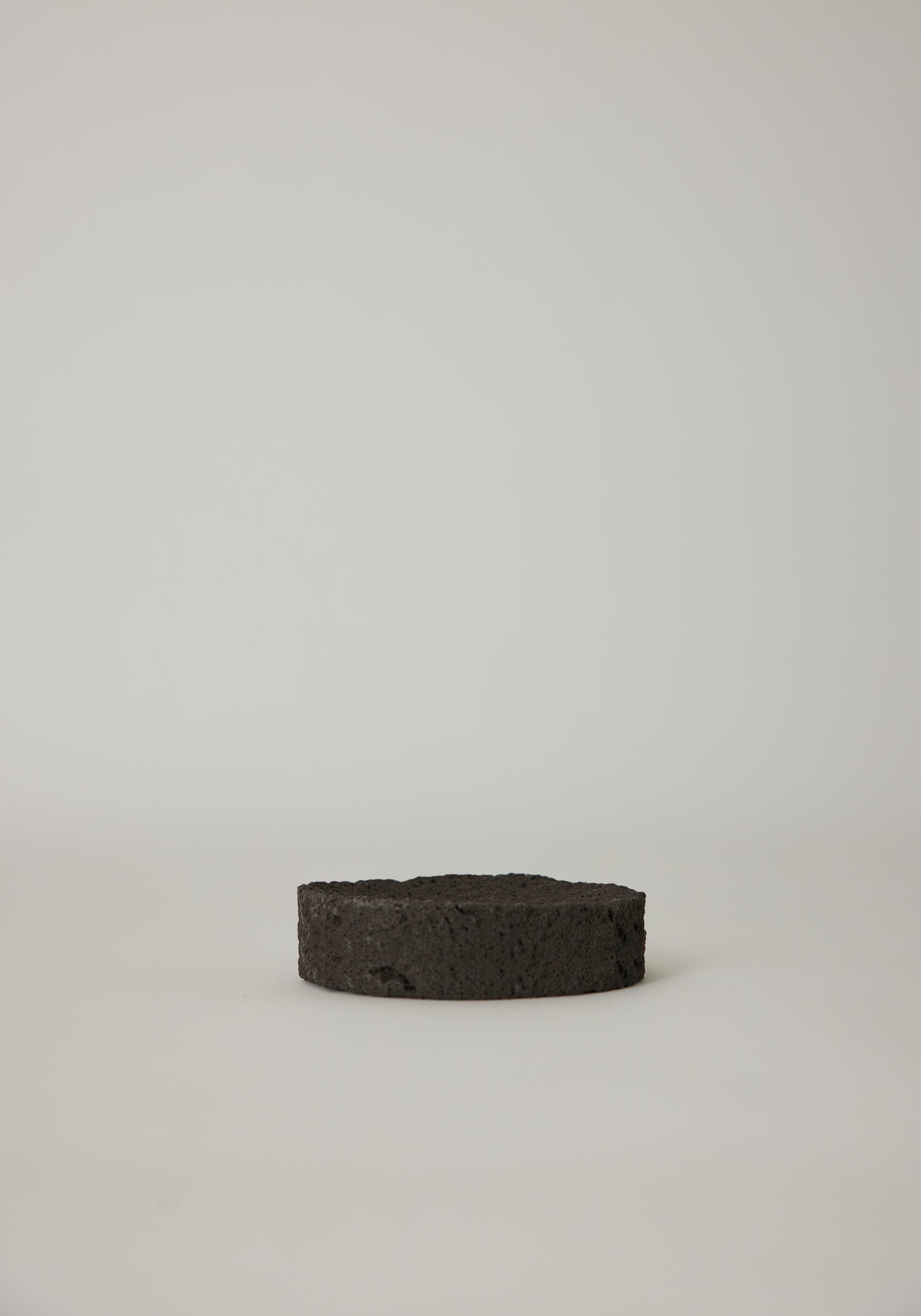 Round Tray made from black, porous lava stone, handcrafted in the Netherlands by Louis Crombag, featuring a flat top and carved indent, measuring 2 cm by 13.5 cm.