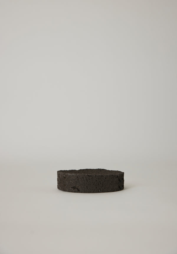 Round Tray made from black, porous lava stone, handcrafted in the Netherlands by Louis Crombag, featuring a flat top and carved indent, measuring 2 cm by 13.5 cm.