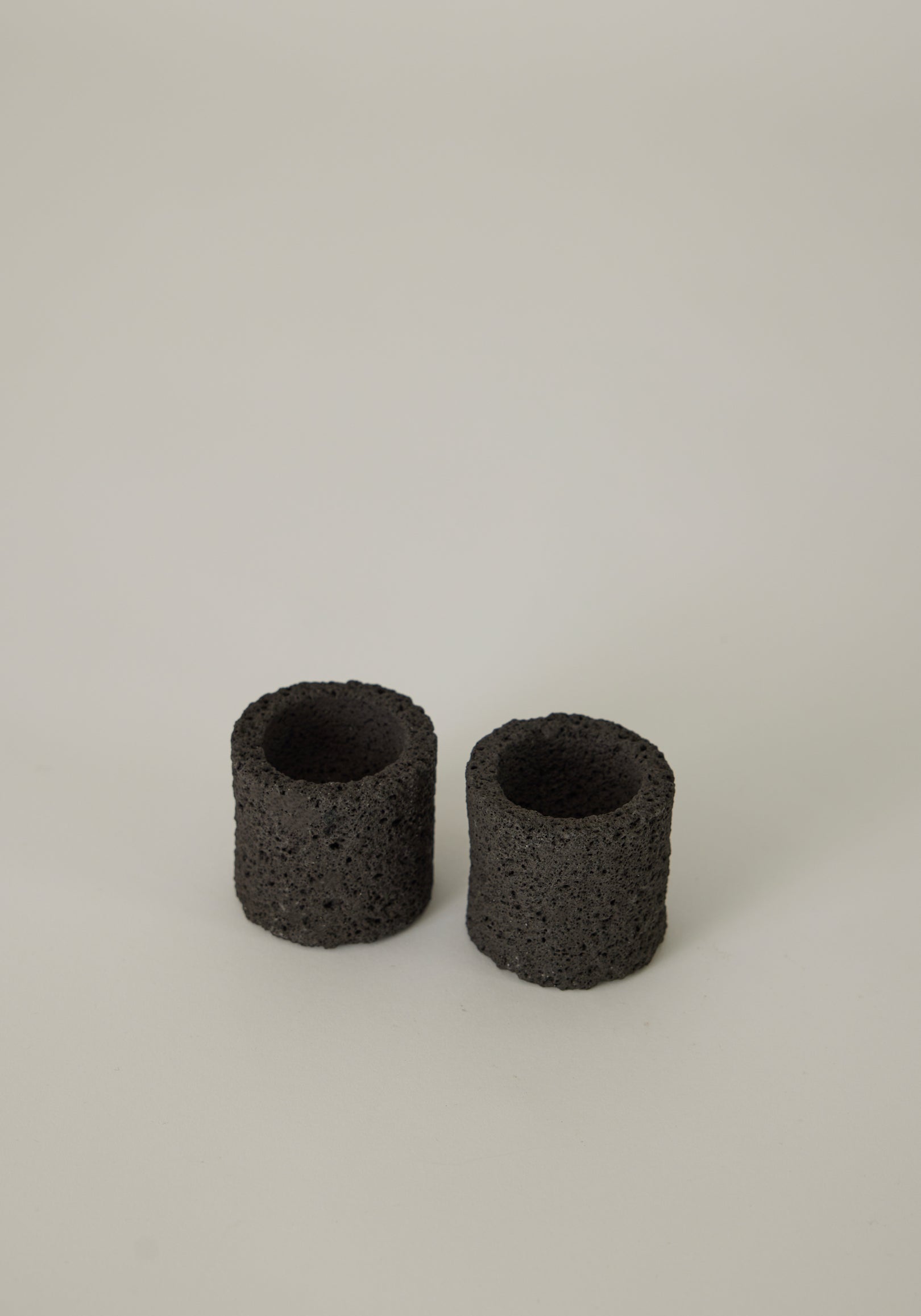 Two handcrafted Egg Cups made from dark, porous lava stone, showcasing a unique, cylindrical design, created by Dutch artist Louis Crombag.