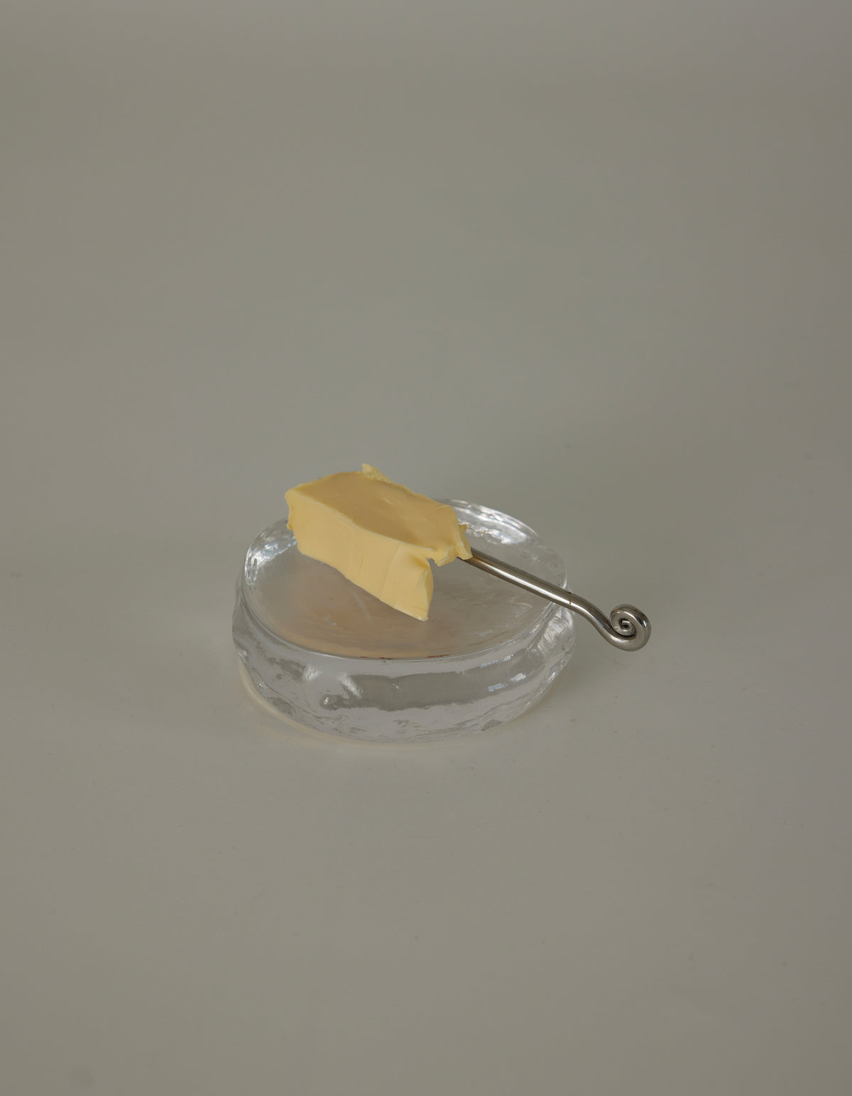 Butter plate | Glass: A clear glass dish holds a rectangular pat of butter, accompanied by a slender silver spreader with a decorative spiral curl handle.