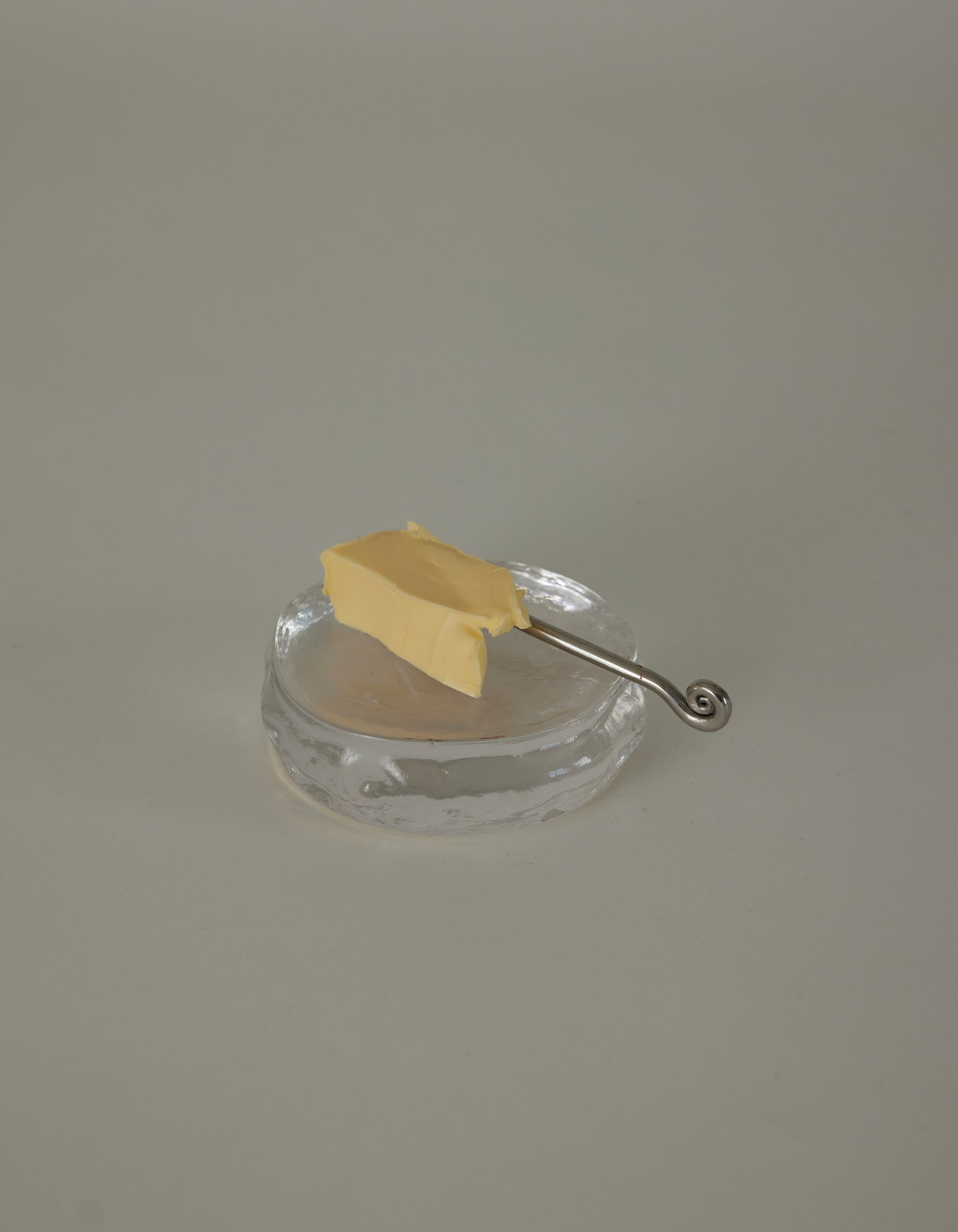 Butter plate | Glass: A clear glass dish holds a rectangular pat of butter, accompanied by a slender silver spreader with a decorative spiral curl handle.