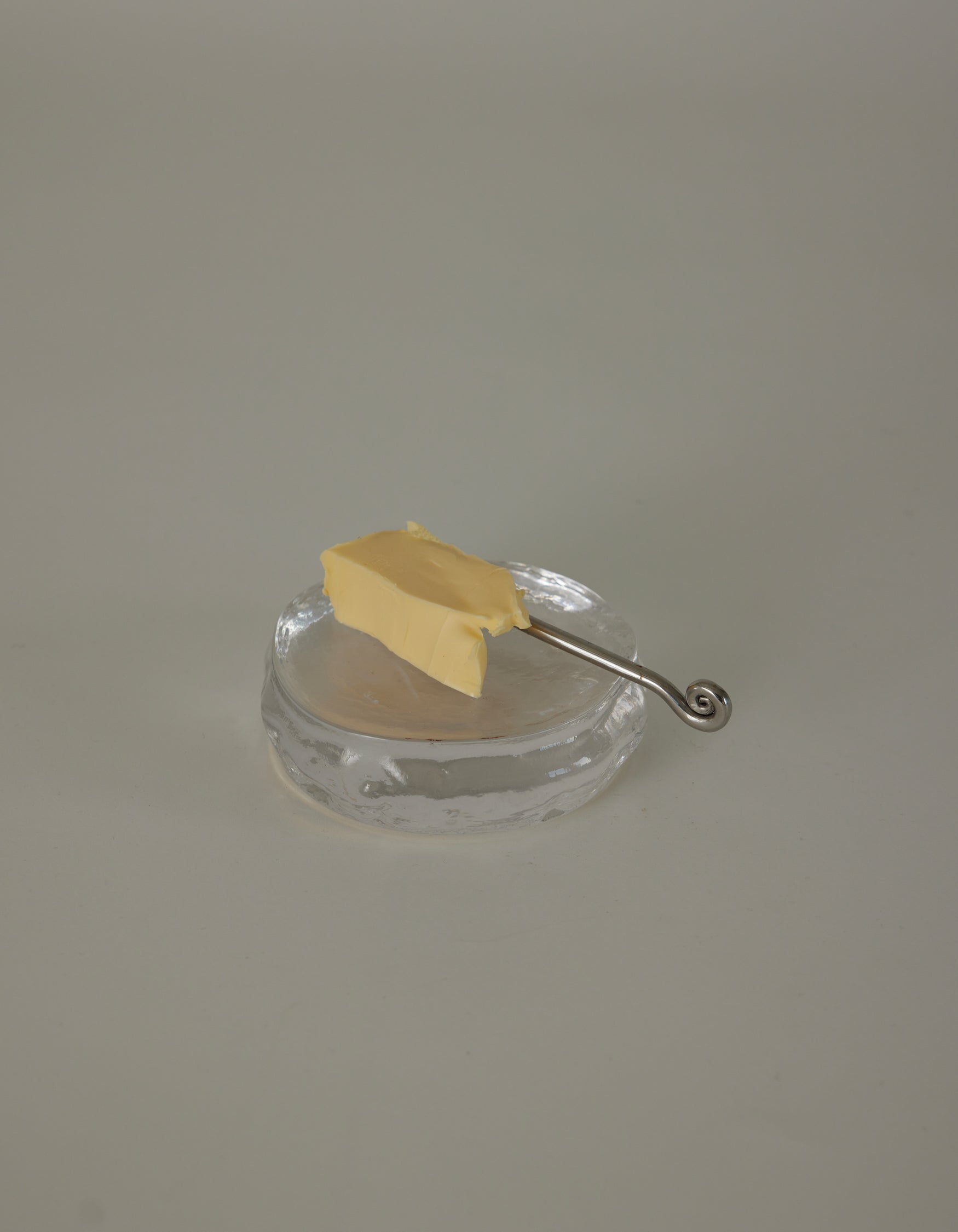 Butter plate | Glass: A clear glass dish holds a rectangular pat of butter, accompanied by a slender silver spreader with a decorative spiral curl handle.
