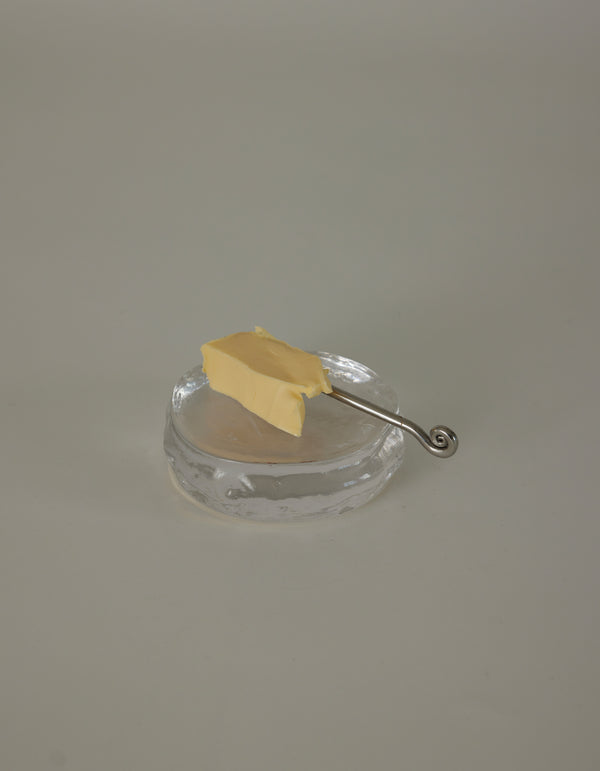 Butter plate | Glass: A clear glass dish holds a rectangular pat of butter, accompanied by a slender silver spreader with a decorative spiral curl handle.