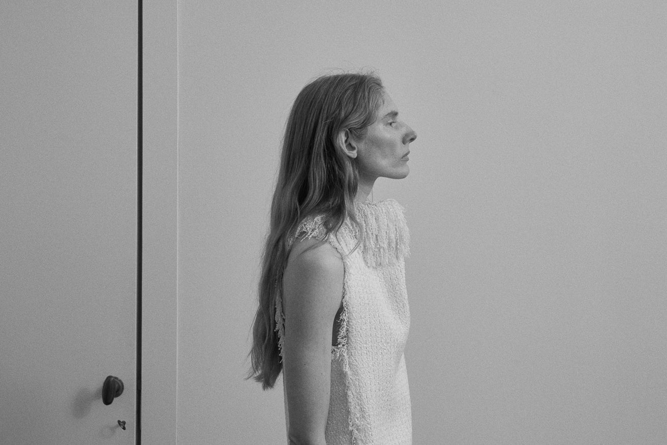 A black and white portrait of a woman in profile wearing a textured sleeveless top, standing against a plain wall next to a door.