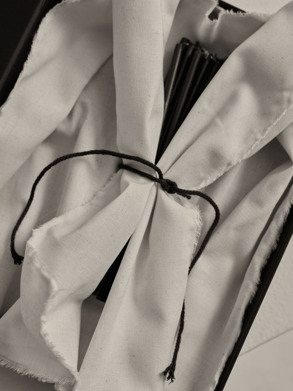 Candles 10mm | noir: A set of 10 slender beeswax candles wrapped in off-white cotton, secured with a black cord, in a shallow tray.