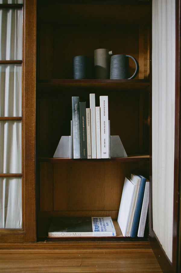Stainless steel bookends holding books upright on a wooden shelf, showcasing their minimalist design crafted from 8mm thick stainless steel in collaboration with Paul Coenen.