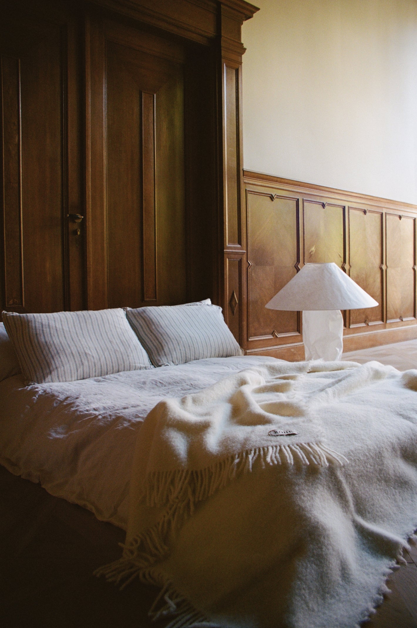 Cream wool blanket with chunky fringes drapes over a bed on a herringbone floor, enhancing a minimalist bedroom with striped pillows and a sculptural lamp.