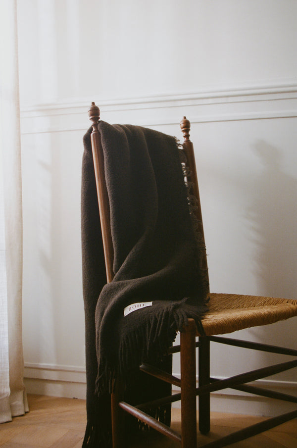 Wool blanket | chocolate draped over a rustic wooden chair, showcasing its chunky fringes and soft texture. Label reads “ROHE”; woven from Italian yarn.