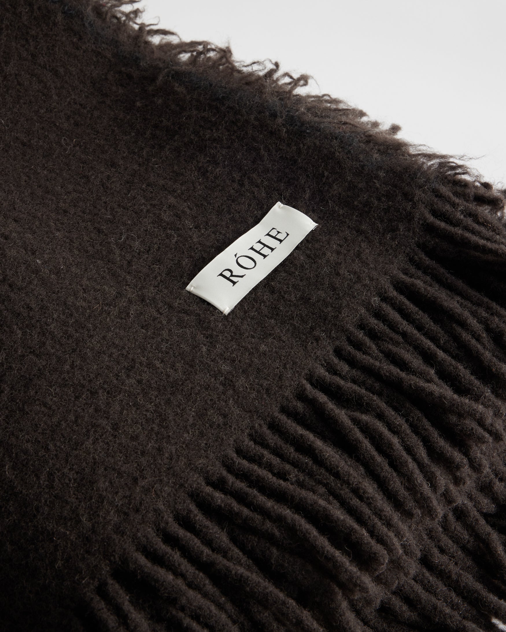 Close-up of a soft wool blanket | chocolate color with fringed edges and a small woven label sewn onto the fabric.