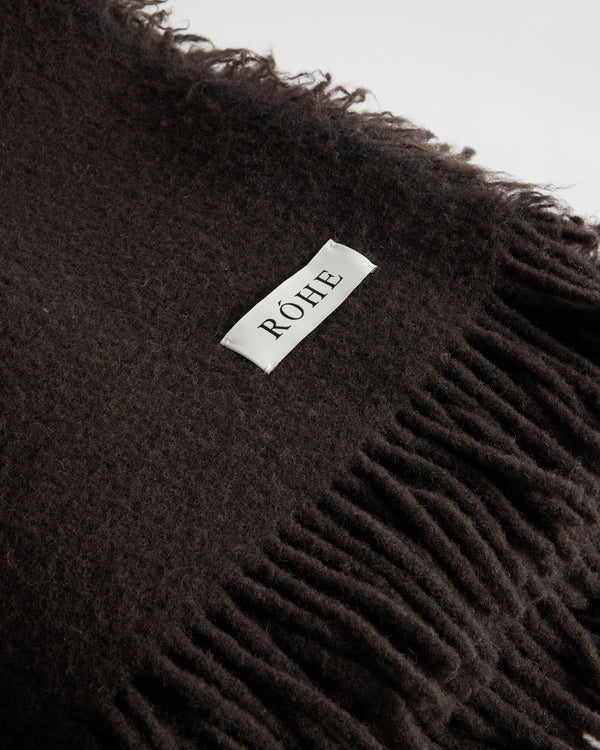 Close-up of the Wool blanket | chocolate, showcasing its dark, fringed woolen texture and a white label with “RÓHE,” made from Italian yarn, 130cm x 180cm.