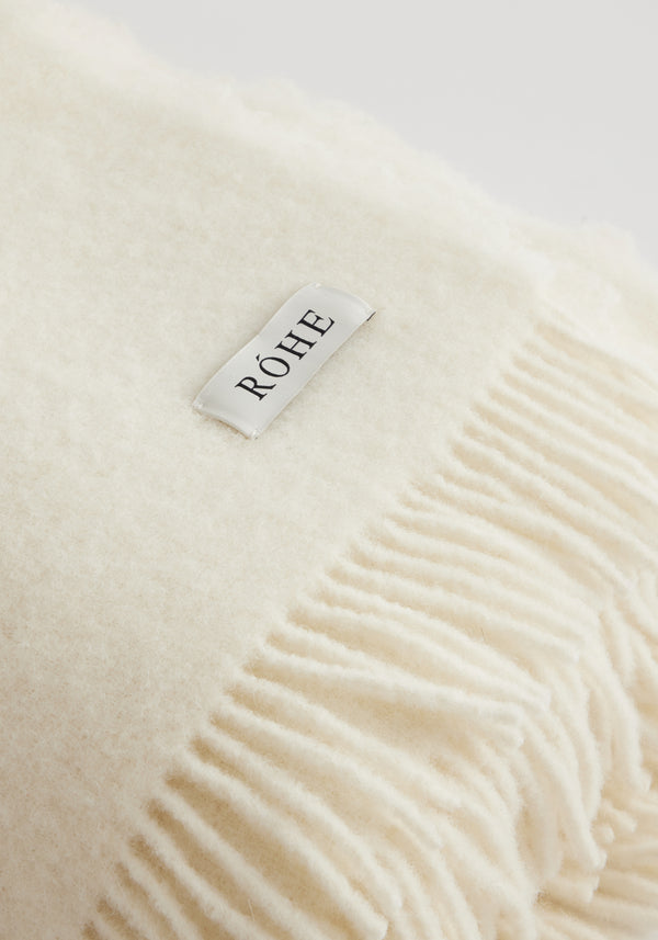 Close-up of the Wool blanket | Cream, showcasing its ivory color, fringed edges, and RÖHE label. Made from Italian virgin wool and polyamide blend.