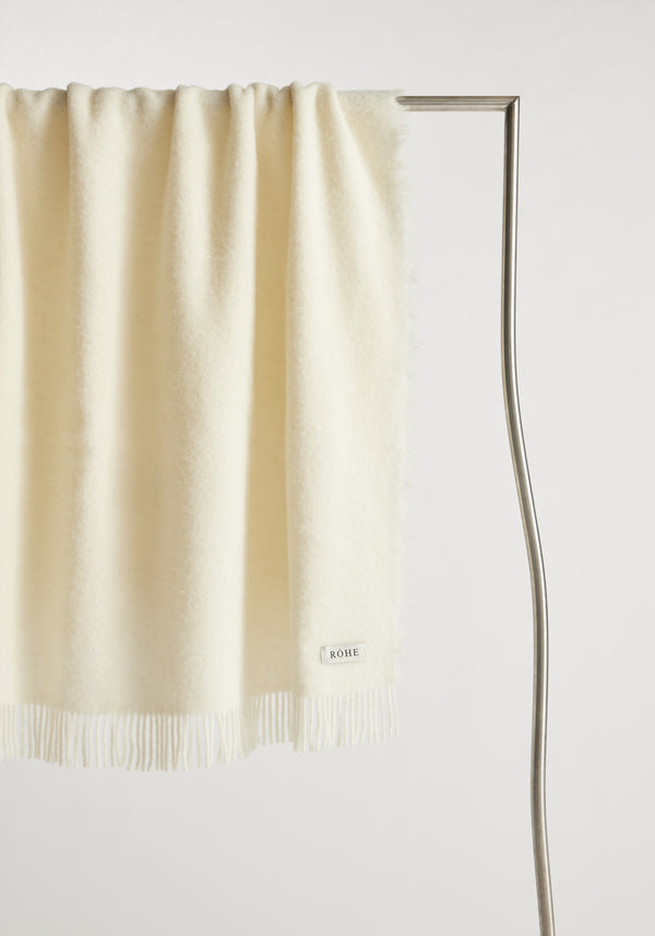 Cream-colored Wool blanket with chunky fringes, draped over a metal rack, features a RÖHE label. Made from Italian virgin wool and polyamide blend.