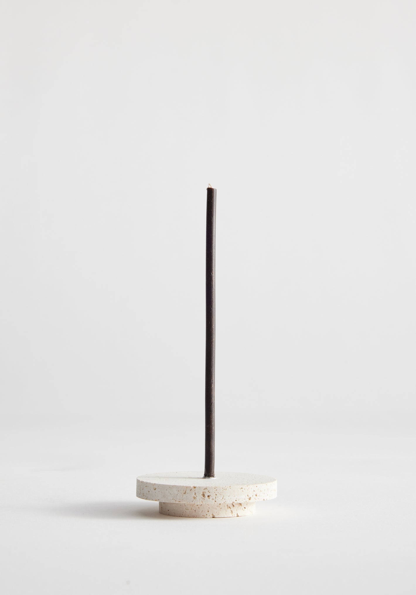 Dish candleholder small | travertine: A single black incense stick stands in a handcrafted round travertine holder by Louis Crombag, showcasing unique craftsmanship.