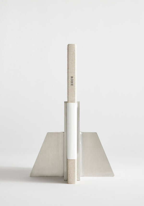 Stainless steel bookends, handcrafted by Paul Coenen, hold a cloth-bound book upright. The minimalist design features angled plates and a vertical support, showcasing Dutch craftsmanship.