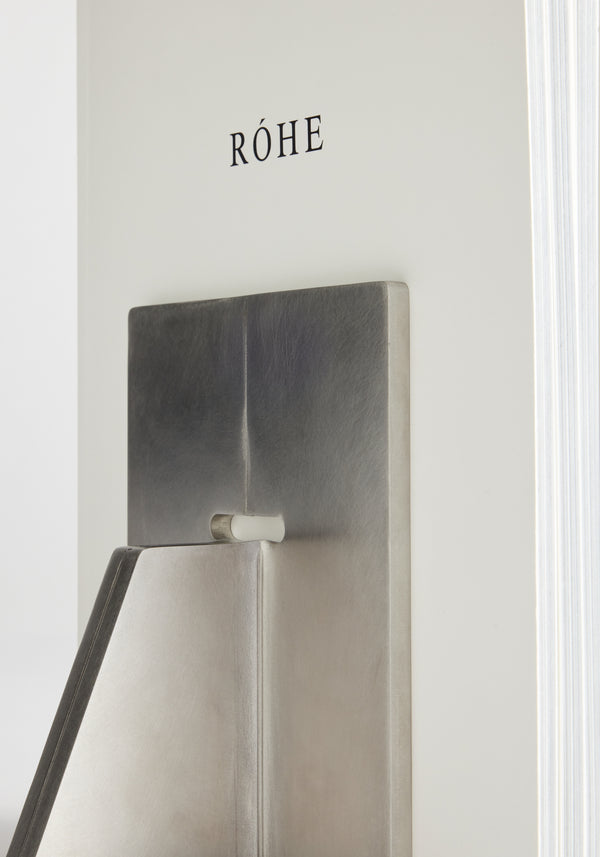 Close-up of a thick book labeled RÓHE with a sculptural stainless steel bookend by Paul Coenen, showcasing its geometric design and craftsmanship.