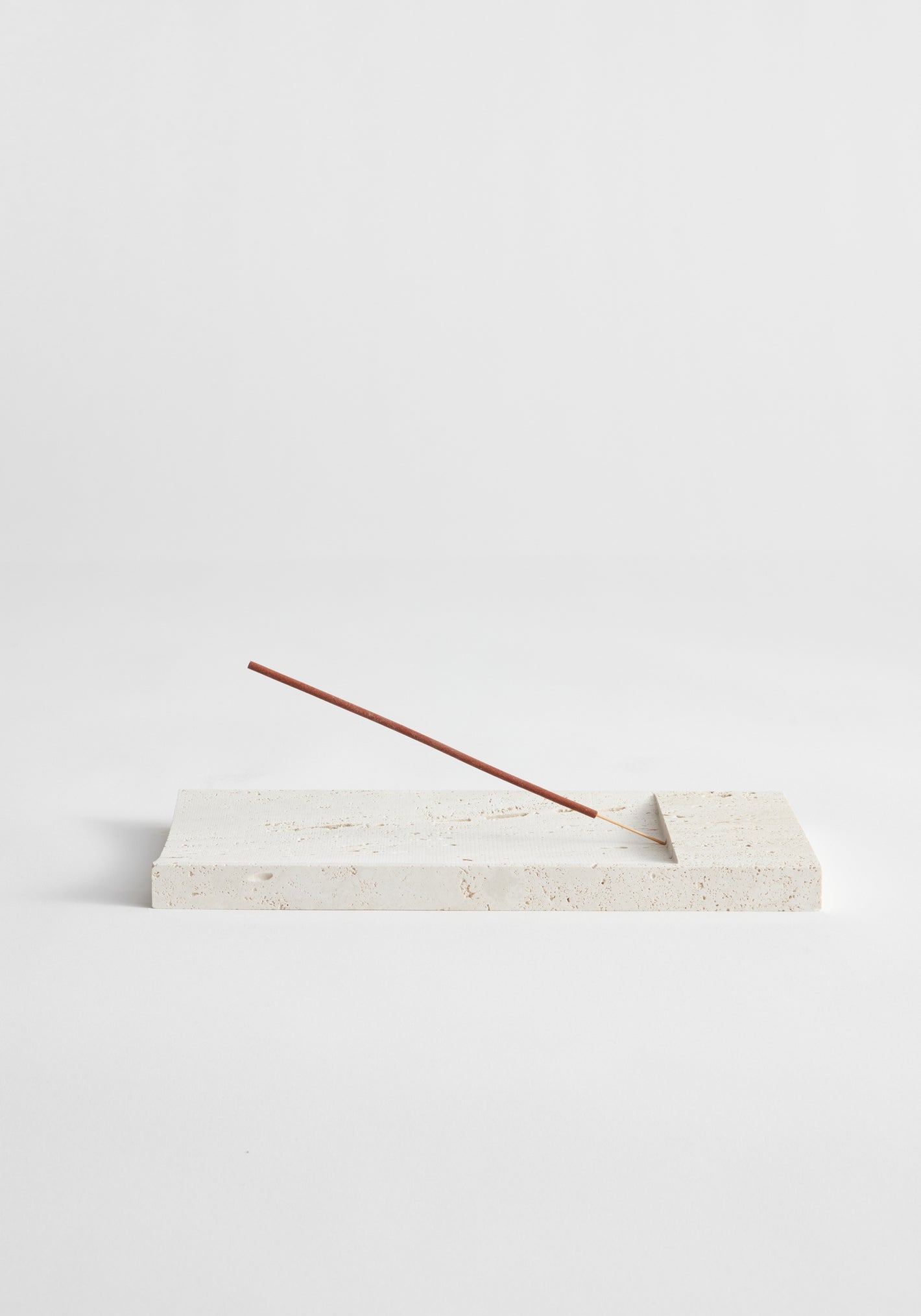 Incense holder tray | travertine: Slender incense stick rests on a handcrafted cream-colored rectangular stone tray, made from leftover Italian travertine by Dutch artist Louis Crombag.