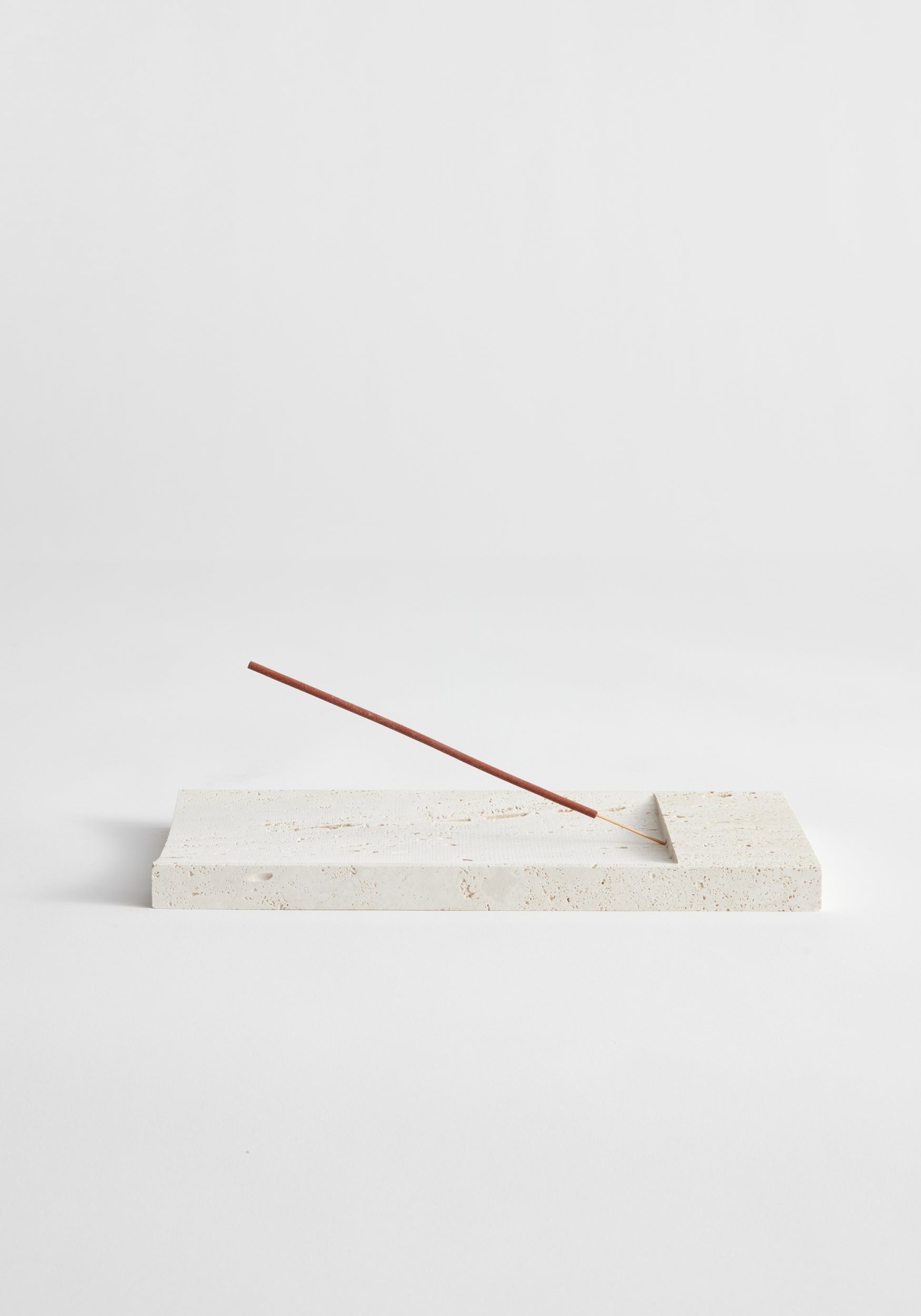 Incense holder tray | travertine: Slender incense stick rests on a handcrafted cream-colored rectangular stone tray, made from leftover Italian travertine by Dutch artist Louis Crombag.