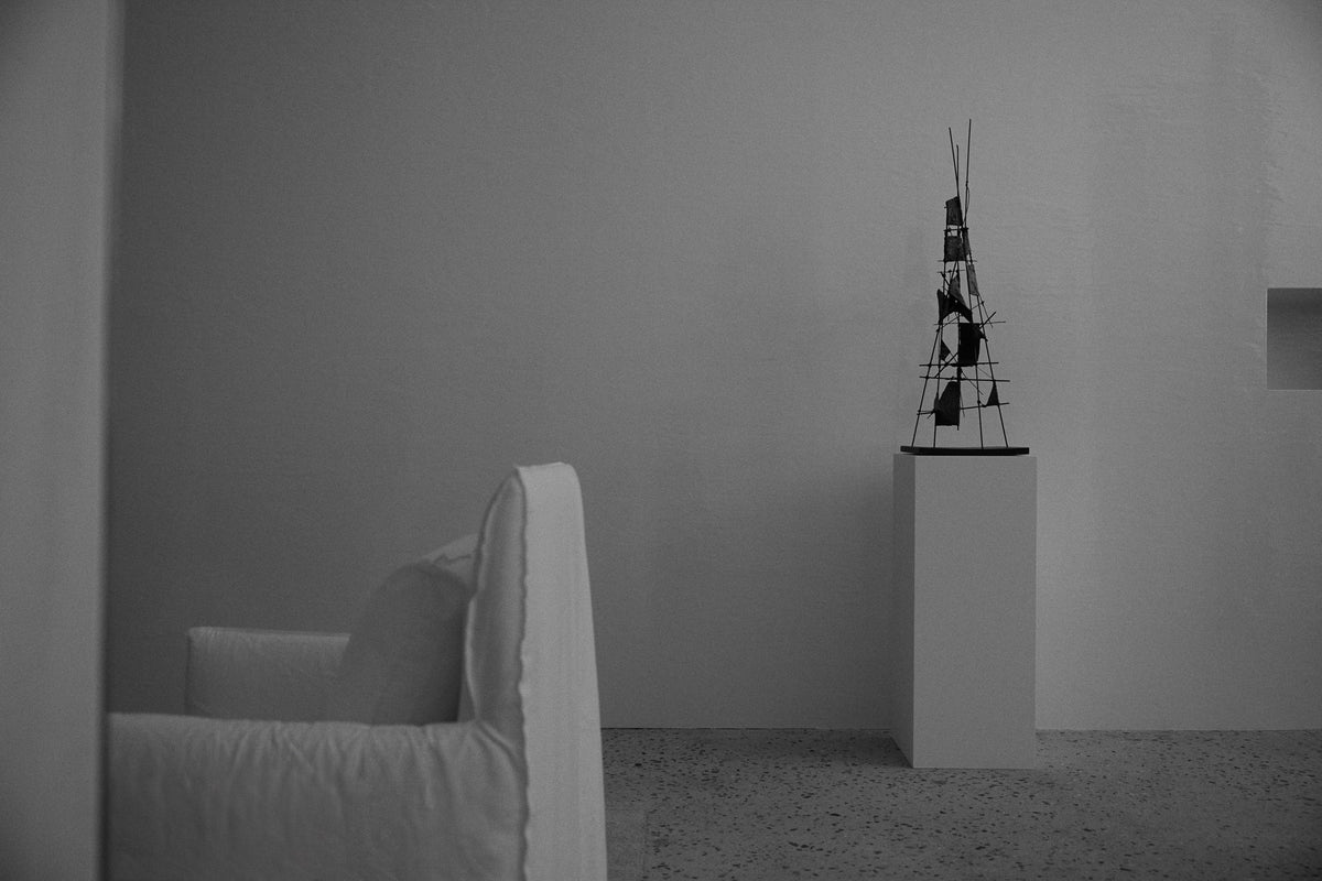 Grayscale minimalist interior with a white upholstered chair in the foreground and an abstract sculpture displayed on a pedestal against a plain wall