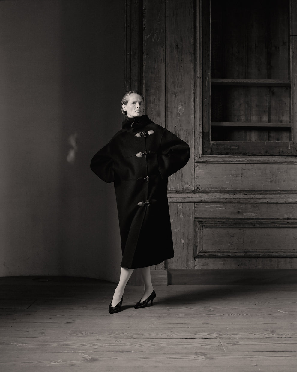 Black and white portrait of a woman standing in a rustic wooden room, wearing a long dark coat with a high collar and pointed heels.
