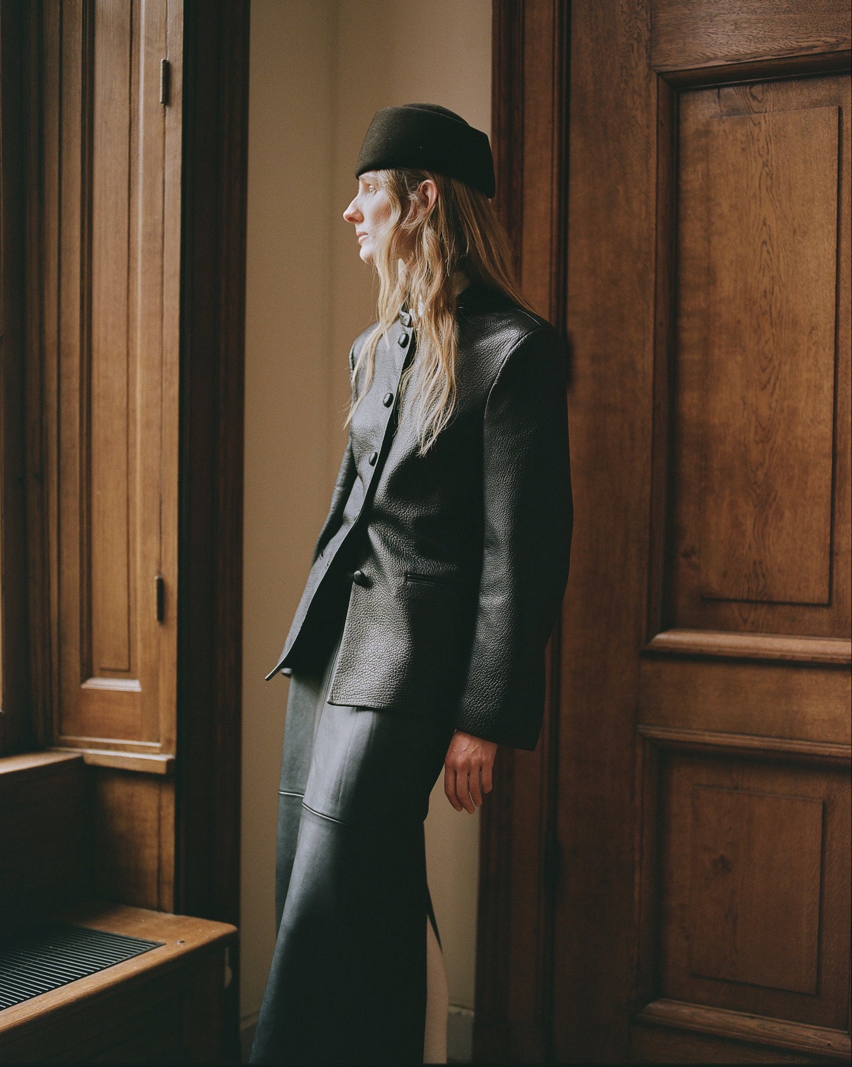 A woman in a dark charcoal textured leather jacket and black pill box hat stands in profile in a doorway with warm wood paneling.