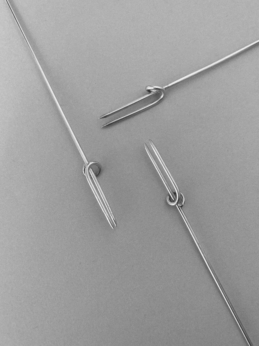 Three slender silver vine forks with double prongs arranged on a gray surface, designed for use in vine training or gardening tasks.